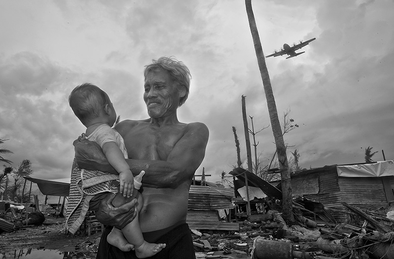 This image shows the quite happy moment in which the rescue planes started to arrive, delivering humanitarian aid sometimes parachuted from the air and beginning to displace the survivors from Tacloban to Cebu City or to Manila.  Remaining as the only two survivors of a family, a grandfather had remained very attached to his granddaughter protecting her throughout the ordeal and as a result of the humanitarian aid he had been able to feed her with some powder milk. Despite that the grandfather had been affected by some minor scratches, the health of both of them was stable. One of the most shocking experiences to observe during the aftermath of the typhoon, was the reaction of the people. Many of them , instead of showing any signs of distress, quite on the opposite they smiled when approached and even laughed when asked about their fate. As I was informed later , that was a way of coping with Post Traumatic Stress Disorder. The magnitude of their losses was so overwhelming that it was just more than a normal human being could bear, so instead of dealing with their sadness at that point they joined other people and tried to keep themselves busy either by cleaning the debris or constructing small in which to live in the short run. As one of the mental health doctors helping them told me, the realistic dimensions of their loss would come to haunt them later, but for the moment it was simply too much.  At some places the children played cheerfully between themselves oblivious to many near cadavers, some of them could have probably been someone from their families.