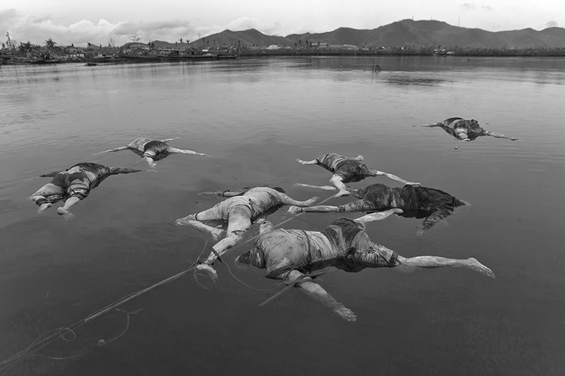 After the devastation created by the rise of the ocean levels, many people drowned at the area of San Jose (Tacloban City), which was one of the areas with most casualties.Improvised teams of fishermen volunteered to gather the corpses at the ocean and had them tied up together and secured by a rope tied to a stone laying at the bottom of the ocean, waiting for the body rescuers to come to pick them up and have them transported to mass graves. Most of them couldnt be recognized either because their relatives and friends were missing as well or because the bodies were beyond recognition. On the other hand the amount of casualties was so high that there was virtually no time for such a procedure (identifying the bodies by relatives or friends) and due to the high risk of spreading epidemics,the final decision was to have them immediately transported to mass graves, although an autopsy center gathered forensic data in an endless daily effort. That day it had been raining mildly and to an extent that there was a certain beauty , silence and calmness at the ocean, and the bodies floated motionless, as if it they had been deposited there on purpose, in an intermediate stage in between life and death and in communication with nature. I approached the bodies while standing on the top and mantaining the equilibrium on the thin canoe of two young fishermen and the sound of the drizzling rain and of their rowing paddles was the only sound to be heard.