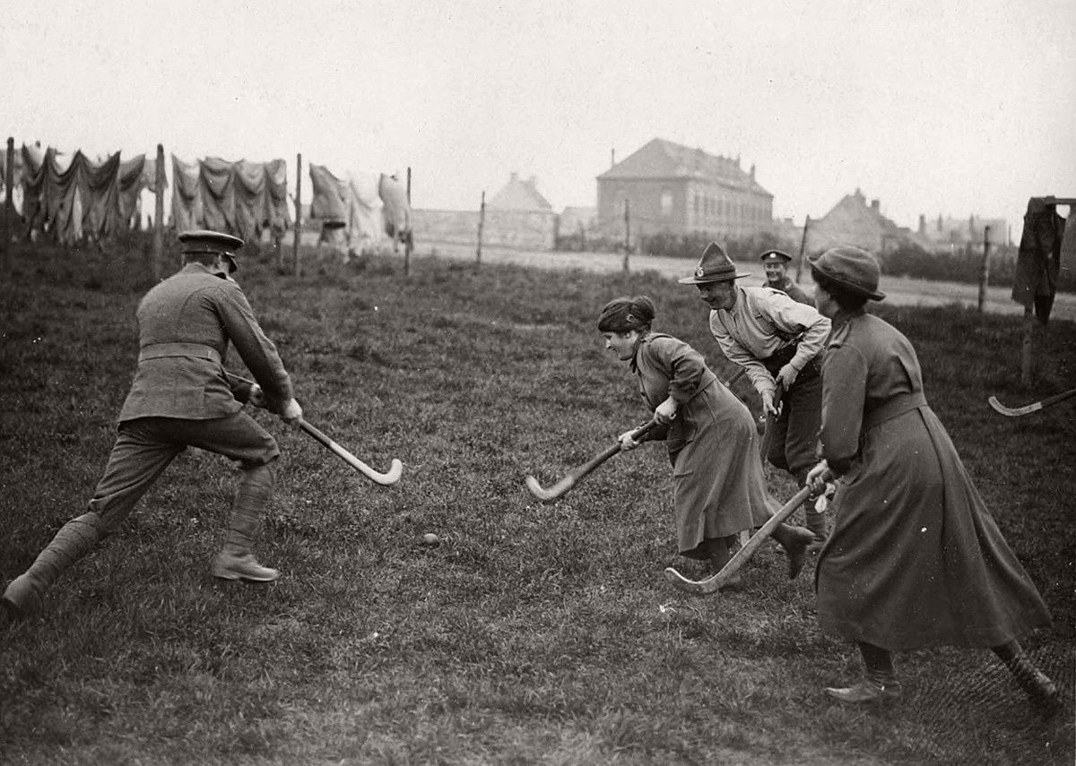   Women's Army Auxiliary Corps (W.A.A.C.) members play field hockey with soldiers in France, during World War I, drying greens and convalescent home buildings visible in the background. # National Library of Scotland