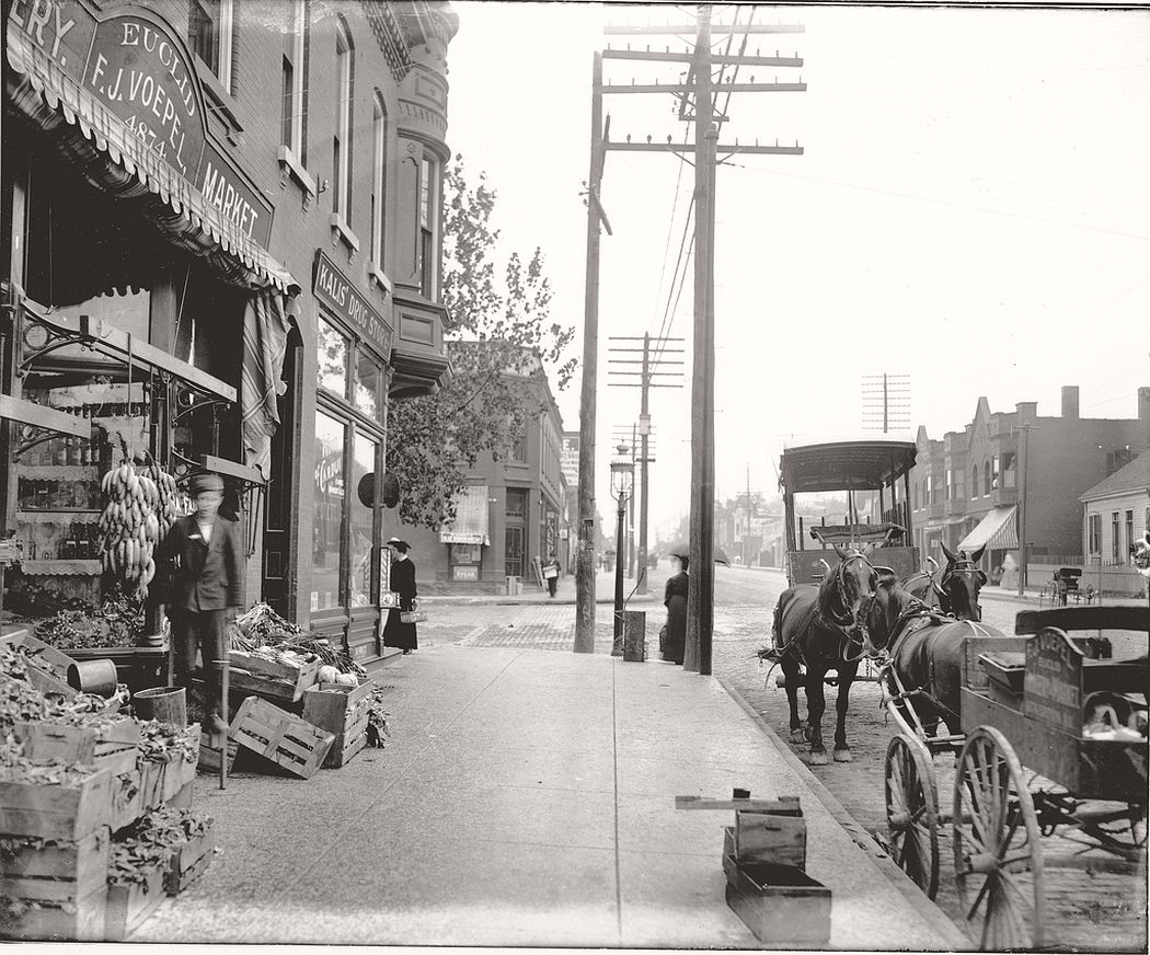 A boy on stilts stands in front of the produce stands in front of the F.J. Voepel Market, 1906