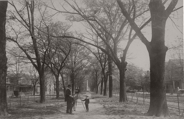 Photographer with view camera in the median of Green Street in Augusta, Georgia.The camera on a tripod is from the dry plate era, late 19th or early 20th century. Two young boys stand with the photographer in a derby in the median of a wide city boulevard lined with large residences. On the right is a carriage with a mounting block and on the left a carriage with a horse. There is a monument in the distance and there appear to be electric light strung across the median.
