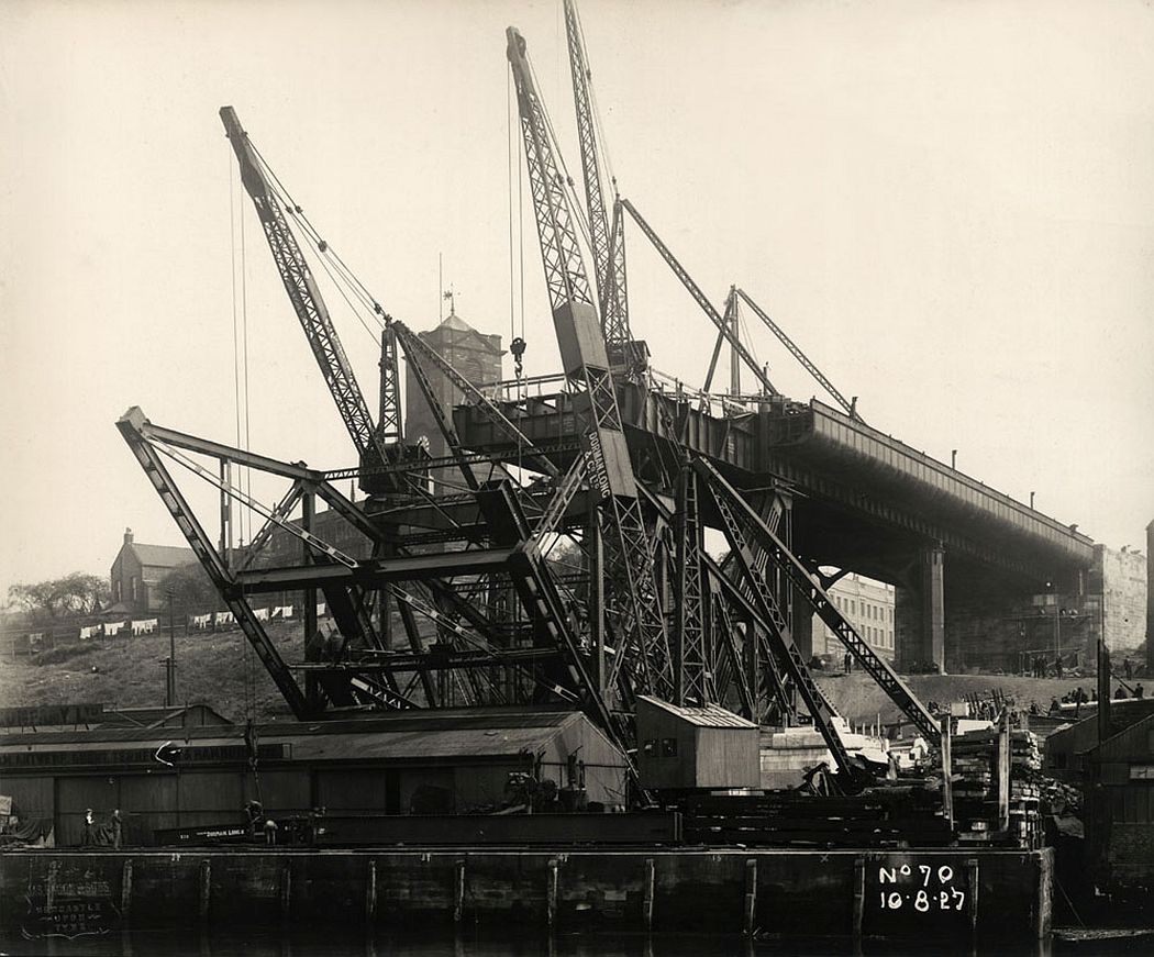The first sections of steelwork rise over Hillgate Quay, Gateshead, 10 August 1927. Most of the girders in this photograph are part of a temporary cradle used to support the first three sections of the bridge's main arch. 