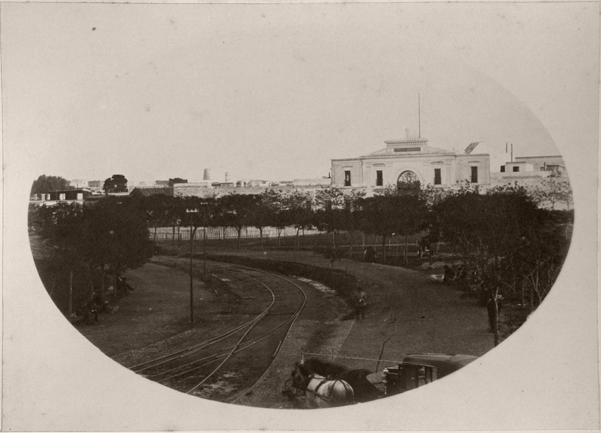 Parque de Artillería Park with its seats, trees and gaslight columns. The photo was taken from the Buenos Aires Western Railway terminus. Rail tracks, a carriage and its horse and the entering to Cuartel de Artillería can be seen in the image.