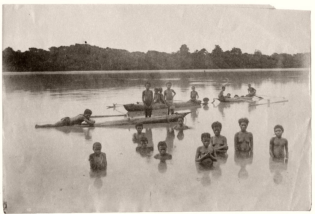 Photograph shows a group of men, women and children standing in a lagoon and on outrigger canoes. Fiji, early 20th century.