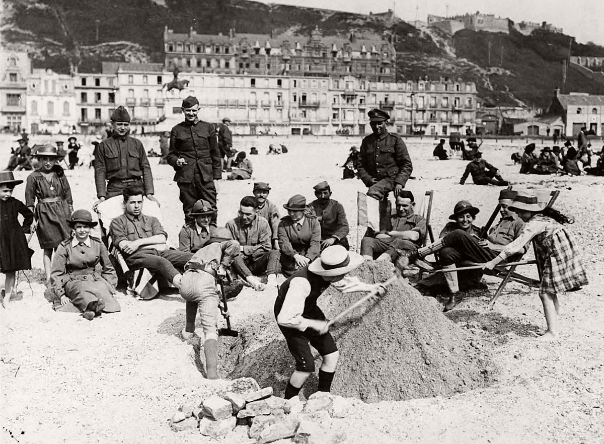   During downtime, soldiers from Britain, France and the USA, plus some members of the Women's Auxiliary Army Corps (WAAC) watch French children playing in the sand, in France, during World War I. # National Library of Scotland