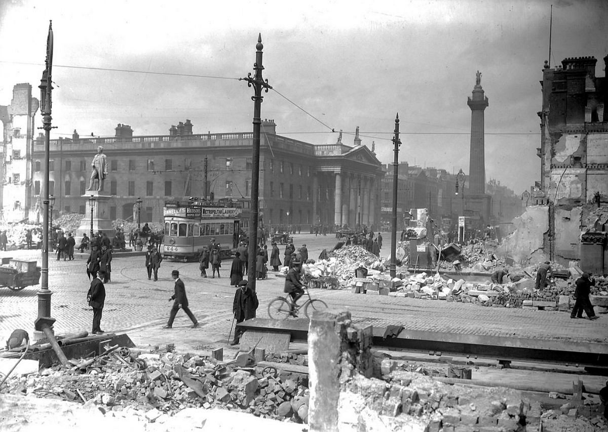 Abbey Street and Sackville Street (O'Connell Street) shelled, rubble remains. The tram passing by was numbered 244. The ads on the tram are for Donnelly's Bacon, Hudson's Super Soap and the Metropolitan Laundry. An ad for Bovil can just be made out on another tram. In the foreground, the bearded man (very nautical vibe) is considering a huge slab on the ground.