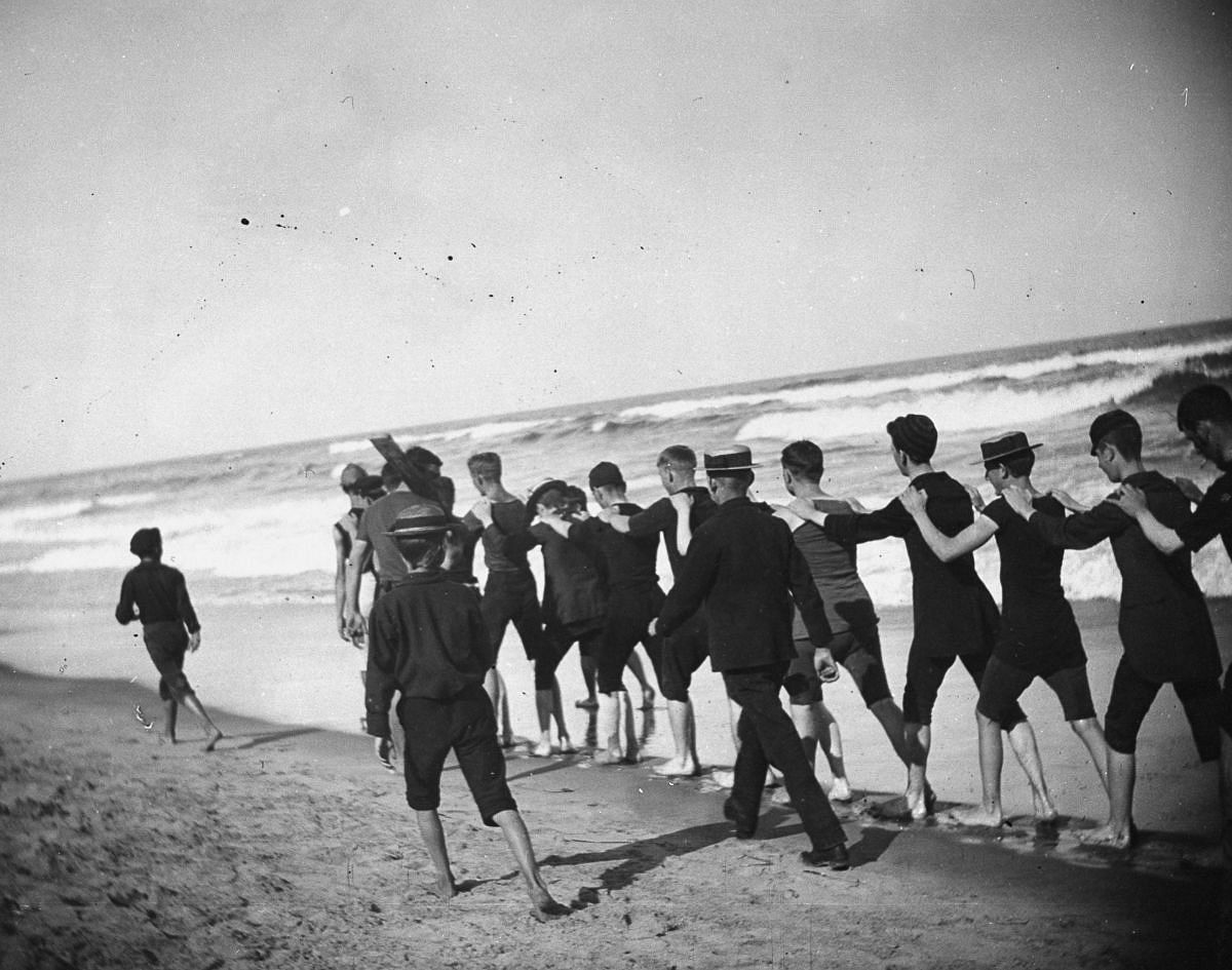 Aug. 23, 1886 - A group of young male bathers walk single file along the beach.