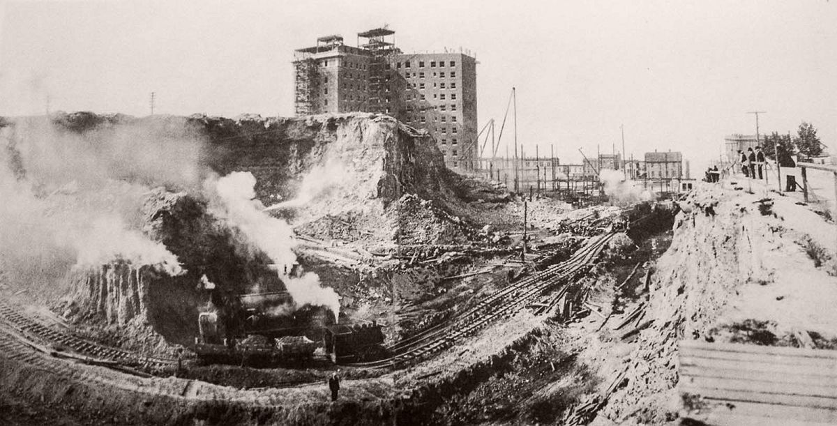 Trains move loose earth at the south summit of Denny Hill near the under-construction New Washington Hotel, at the corner of Second Avenue and Stewart Street. 1907.